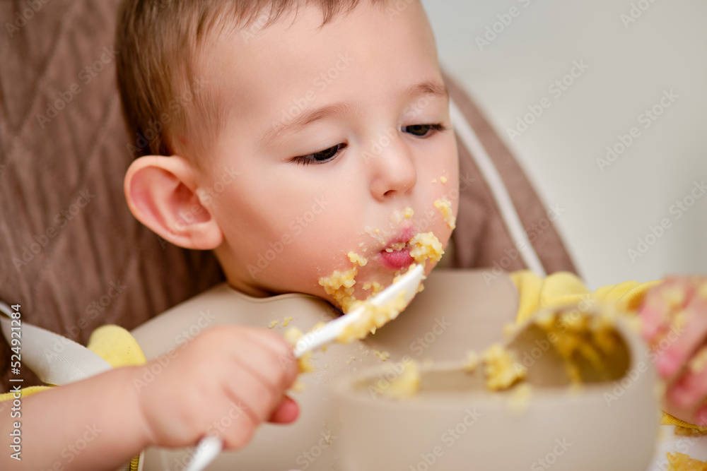 Happy toddler baby boy learns to eat porridge himself with a spoon while sitting in a child chair. Smiling child eats with his hands and a spoon from a plate, kid aged one year Happy toddler baby boy learns to eat porridge himself with a spoon while sitting in a child chair. Smiling child eats with his hands and a spoon from a plate, kid aged one year