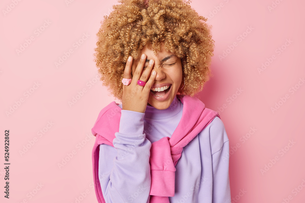 Happy curly teenage girl makes face palm laughs gladfully dressed in casual jumper and pullover tied over shoulders laughs gladfully has fun hears something funny isolated over pink background. Happy curly teenage girl makes face palm laughs gladfully dressed in casual jumper and pullover tied over shoulders laughs gladfully has fun hears something funny isolated over pink background.