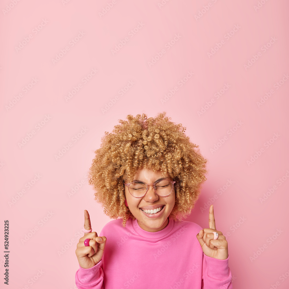 Overjoyed curly haired woman breaks news on top points index fingers upwards shows awesome advertisement wears big spectacles and pullover isolated over pink background. People and promotion
