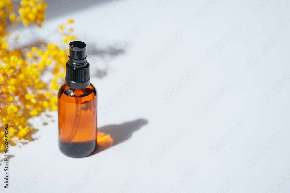 Amber pump bottle with serum, tonic or essential oil with yellow flowers. White background with daylight. Beauty concept for face and body care Amber pump bottle with serum, tonic or essential oil with yellow flowers. White background with daylight. Beauty concept for face and body care