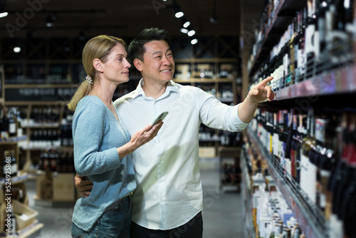 Diverse young married couple man and woman shoppers in supermarket, choosing alcohol wine, using app on phone to identify and scan wine bottle products