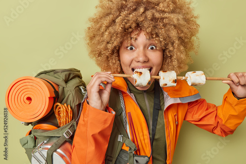 Photo of active female hiker has delicious snack during picnic bites delicious roasted marshmallow carries backpack with equipment spends free time hiking in mountains poses indoor. Adventure #524926038