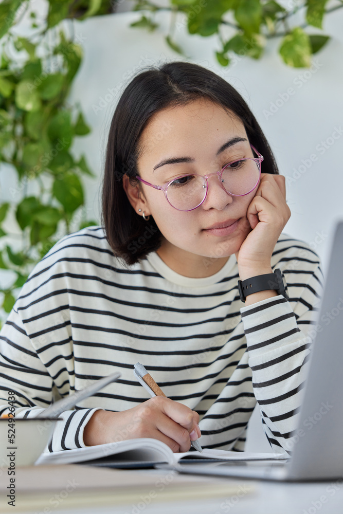 Pretty Asian woman works online on laptop computer sits in coworking space wears transparent eyeglasses and striped jumper writes notes in notebook browses website on laptop computer watches webinar Pretty Asian woman works online on laptop computer sits in coworking space wears transparent eyeglasses and striped jumper writes notes in notebook browses website on laptop computer watches webinar