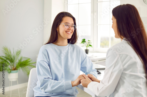 Young female patient rejoices in support of friendly doctor or nurse during medical examination. Professional female doctor tells patient good news by holding her hand. Medicine concept.