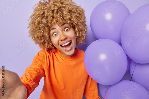 Amazed cheerful woman with curly hair stares bugged eyes keeps mouth opened poses for making photo near bunch of inflated balloons wears orange jumper celebrates anniversary. Party time concept