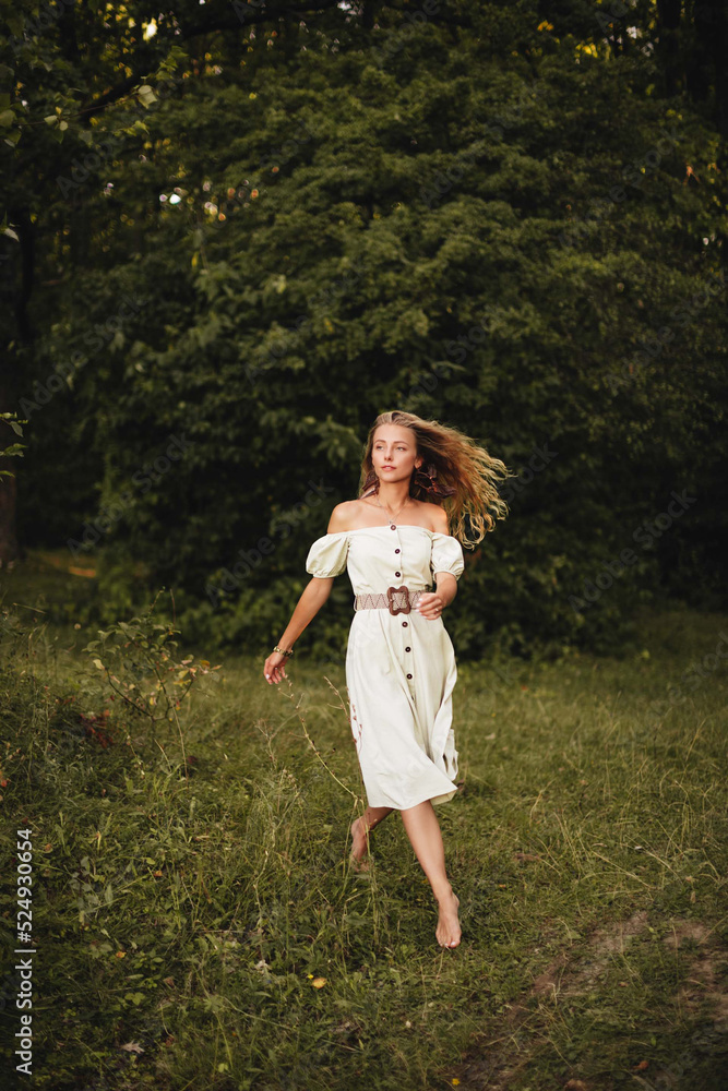 A stylish blonde girl with long curly hair and in a light long dress walks barefoot in the summer forest. A stylish blonde girl with long curly hair and in a light long dress walks barefoot in the summer forest.