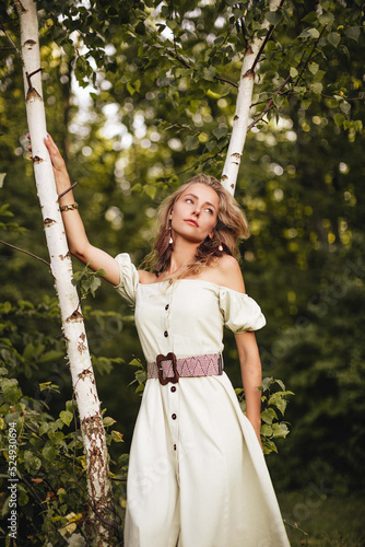 Stylish blonde girl with long curly hair in a light long dress and with earrings with feathers in the summer forest. 