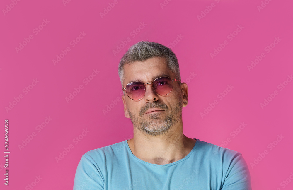 Handsome man 30s 40s wearing pink sunglasses and blue t-shirt with pensive, concentrated look isolated on pink background. Grey haired mature man studio shot Handsome man 30s 40s wearing pink sunglasses and blue t-shirt with pensive, concentrated look isolated on pink background. Grey haired mature man studio shot