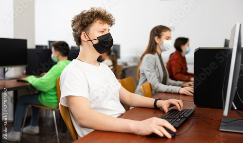 Teenage boy in face mask sitting at table and using computer during lesson. © JackF