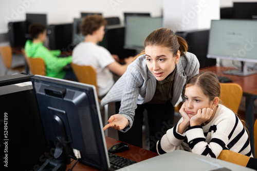 Teacher helping sad young girl to solve computer problem during lesson.