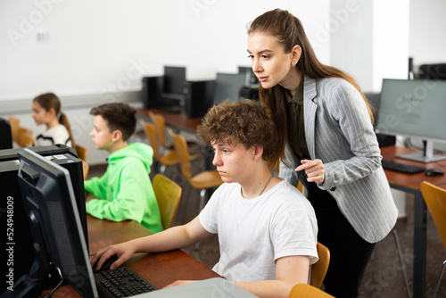 Young female teacher helping focused teenage student studying for exam in college library computer lab..