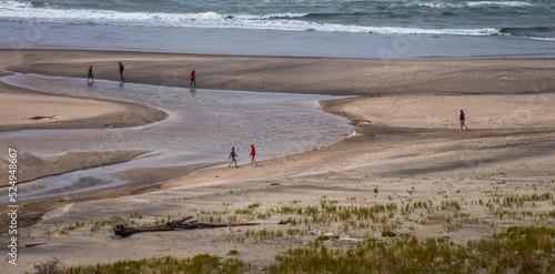 Six people are walking on a beach. The beach has a large inlet of water. One person is wading in that water. Three have red tops and one has a hoody. A surf is breaking at the top. Sea grass is bottom