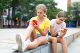 Two young girls sitting in park and using their smartphones.