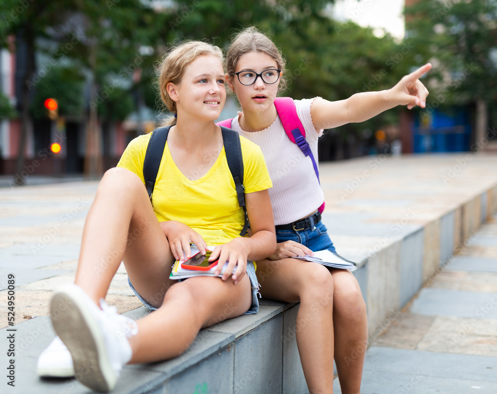 Two friends are sitting on street parapet. One of them points with her hand to something