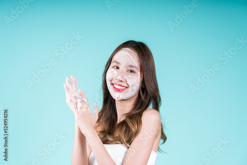 Happy charming young Asian woman applying facial foam cleanser wash and scrub on her face on the blue background.