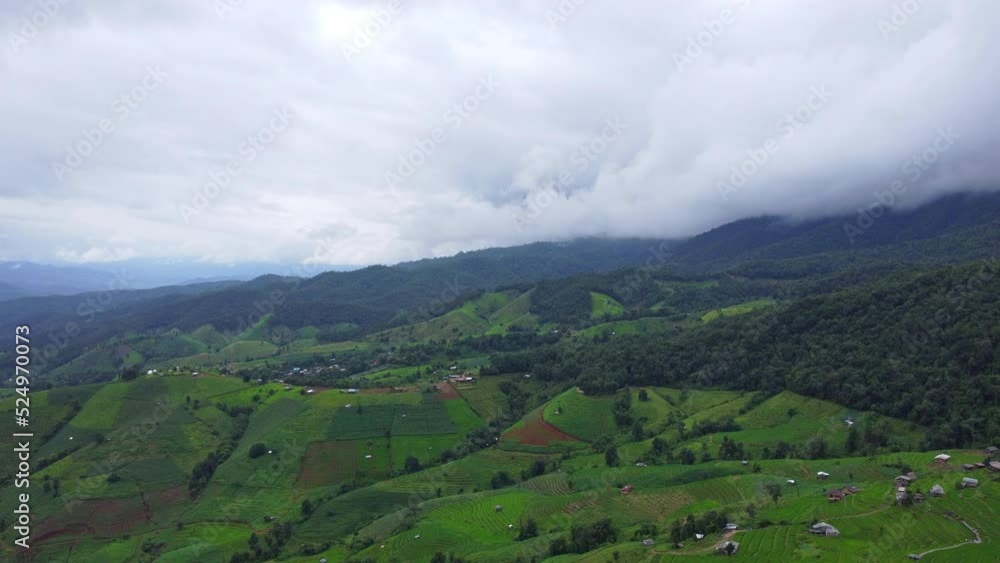 Aerial view of green wave fields with mountains and cloudy sky in rainy season. Beautiful green area of young rice fields or agricultural land in northern Thailand. Beautiful natural landscape backgro Aerial view of green wave fields with mountains and cloudy sky in rainy season. Beautiful green area of young rice fields or agricultural land in northern Thailand. Beautiful natural landscape backgro