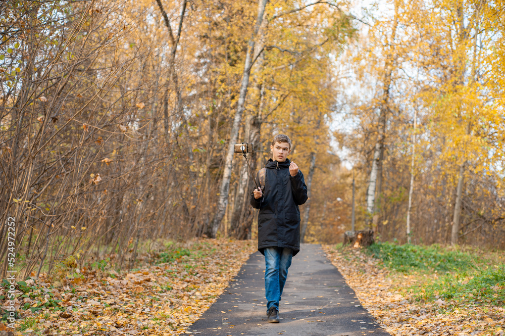 A teenage boy walks through an autumn park and shoots a video. A Caucasian boy in a black jacket holds a monopod with a smartphone. Blogger. Emotional communications.. A teenage boy walks through an autumn park and shoots a video. A Caucasian boy in a black jacket holds a monopod with a smartphone. Blogger. Emotional communications..