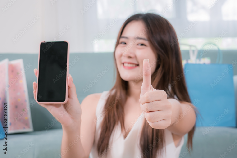 Smiling asian woman present display mobile phone for advertising content her sitting near sofa in living room at home, Shopping online store concept Smiling asian woman present display mobile phone for advertising content her sitting near sofa in living room at home, Shopping online store concept
