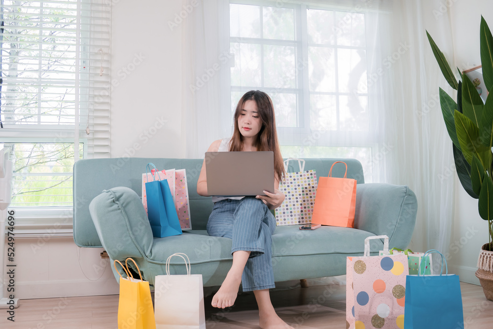 Online shopping concept, Asian woman use laptop computer buying everything on shopping store mall online with shopping bags her sitting on sofa in living room at home Online shopping concept, Asian woman use laptop computer buying everything on shopping store mall online with shopping bags her sitting on sofa in living room at home