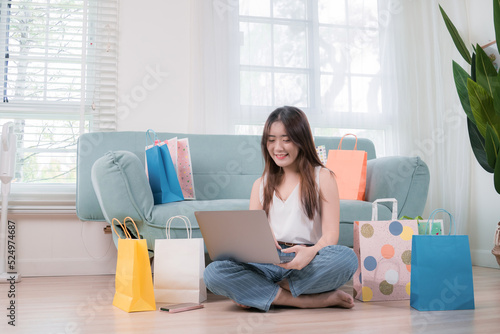 Online shopping concept, Asian woman use laptop computer buying everything on shopping store mall online with shopping bags her sitting on sofa in living room at home