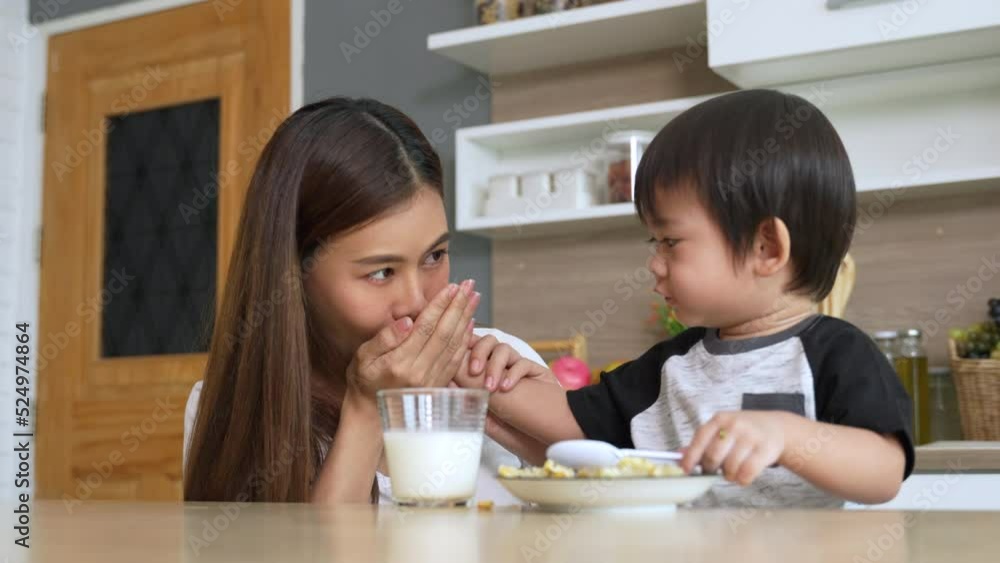 Cute little Asian boy learning to eat by himself and feeding his mother in the kitchen at house. Cute little Asian boy learning to eat by himself and feeding his mother in the kitchen at house.