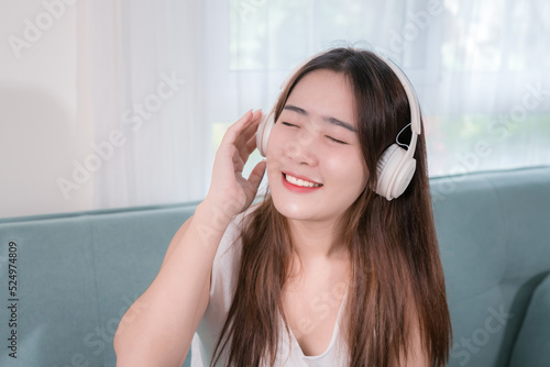 Asian woman use smartphone and wearing headphone listening music entertainment her enjoy and fun sitting on sofa in living room at home