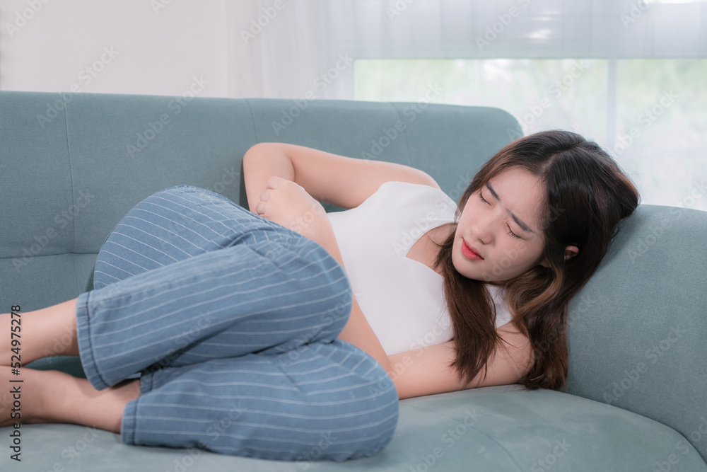 Asian woman with stomachache sitting on sofa in living room at home Asian woman with stomachache sitting on sofa in living room at home