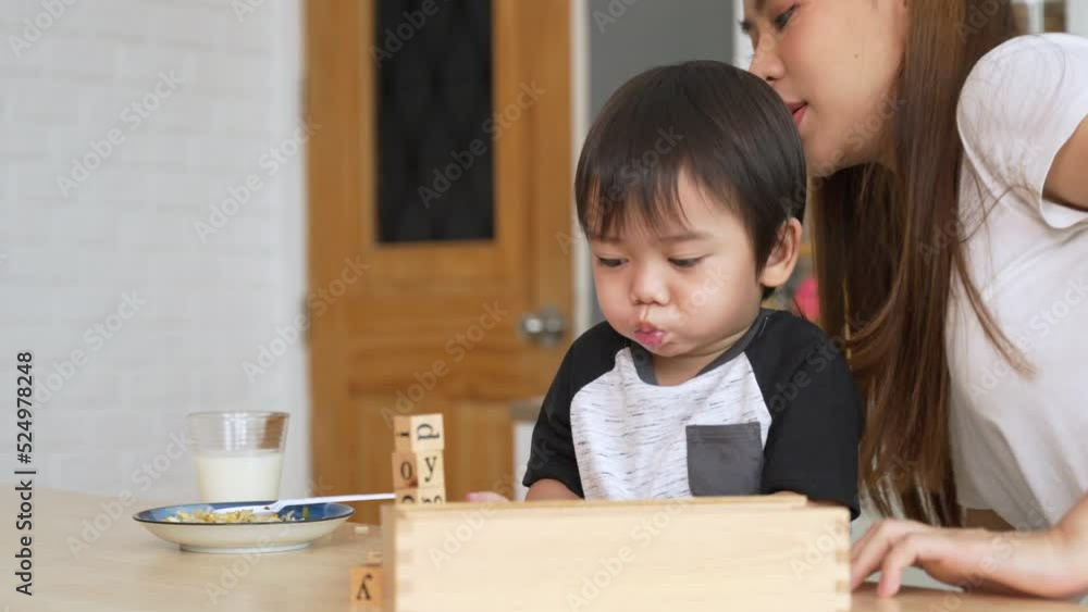Asian young mother is taking her cute little son to play with toys while eating in the kitchen at house. Asian young mother is taking her cute little son to play with toys while eating in the kitchen at house.
