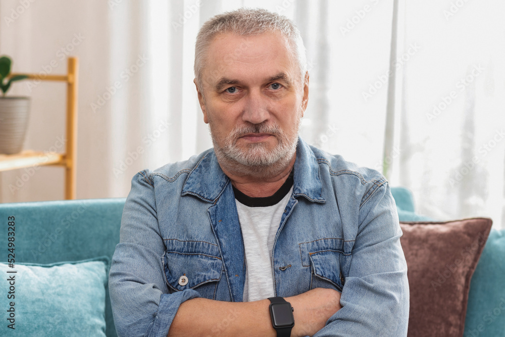 Angry senior man with gray hair and crossed arms looking at camera, sitting on sofa at home Angry senior man with gray hair and crossed arms looking at camera, sitting on sofa at home