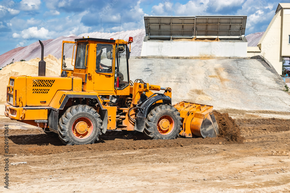 Obraz premium A large front loader pours sand into a pile at a construction site. Transportation of bulk materials. Construction equipment. Bulk cargo transportation. Excavation.