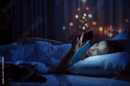 Young woman with her cat using smartphone in bed in her cozy bedroom at night.