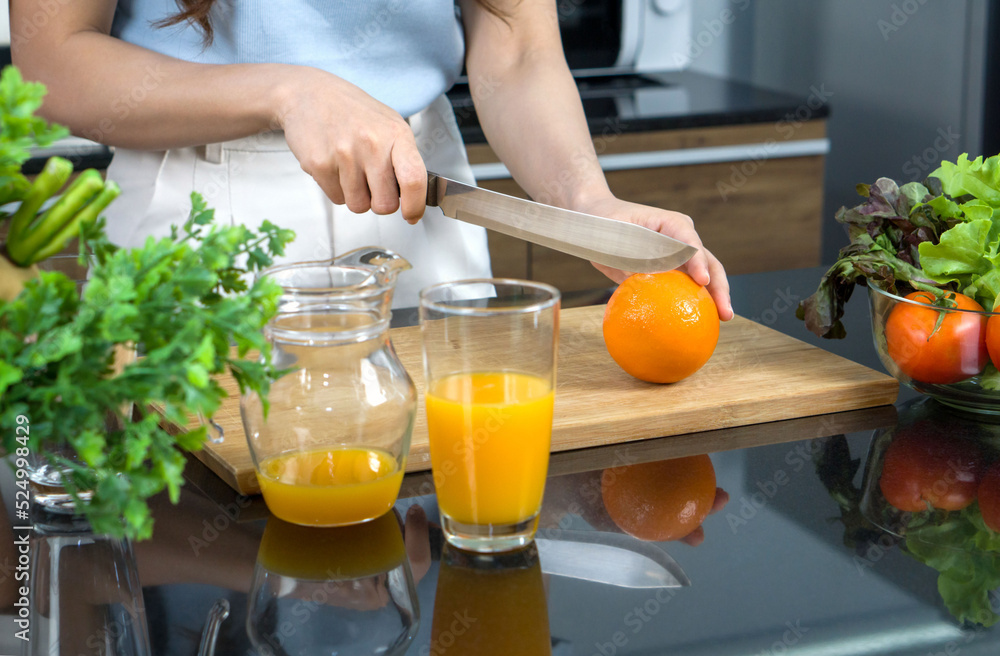 Closeup hand holding knife cutting orange fruit on a wooden chop board. Jar with mixed fruit juice and glass bowl with a variety of fruits and vegetables is placed on the kitchen counter. Closeup hand holding knife cutting orange fruit on a wooden chop board. Jar with mixed fruit juice and glass bowl with a variety of fruits and vegetables is placed on the kitchen counter.