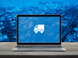 Delivery truck flat icon on modern laptop computer screen on wooden table over city tower, street, expressway and skyscraper, Business transportation online service concept