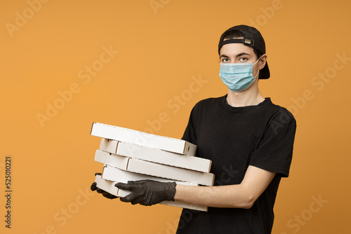 Pizza delivery man, a man in a black t-shirt, cap, protective mask and gloves with boxes in his hand on a yellow background with copy space