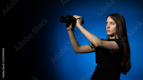 young beautiful woman with long hair in a black t-shirt with binoculars in her hands on a blue background with copy space © Sergey