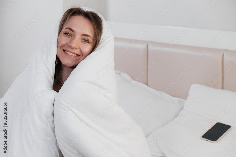 Pretty young smiling Caucasian woman waking up in the morning and stretching in her bed and looking content. Covered in white linen. Pretty young smiling Caucasian woman waking up in the morning and stretching in her bed and looking content. Covered in white linen.