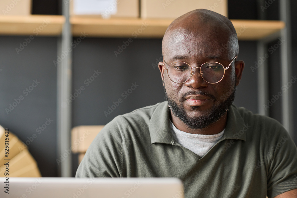 African serious man in eyeglasses working online using laptop in warehouse African serious man in eyeglasses working online using laptop in warehouse