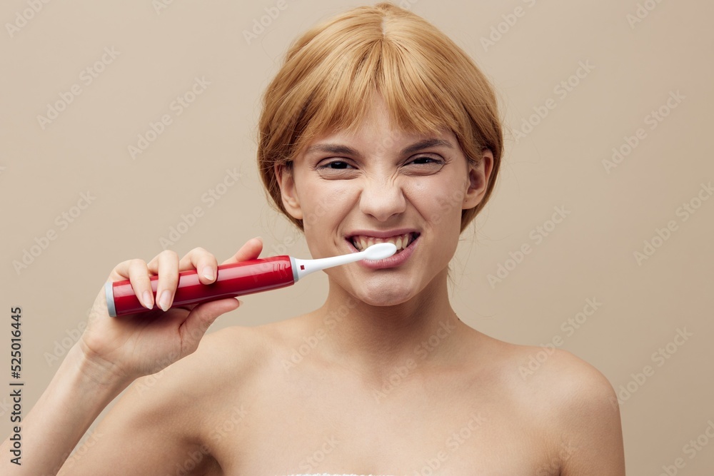 a joyful, funny woman, blonde, with her hair pinned back, stands on a beige background wrapped in a towel and brushes her teeth with a red electric brush, happily squinting her eyes. a joyful, funny woman, blonde, with her hair pinned back, stands on a beige background wrapped in a towel and brushes her teeth with a red electric brush, happily squinting her eyes.