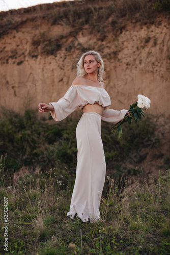 A stylish blonde girl with tanned skin and curly hair stands in a beige suit with a bouquet of flowers on an overgrown quarry. Fashion and lifestyle concept.