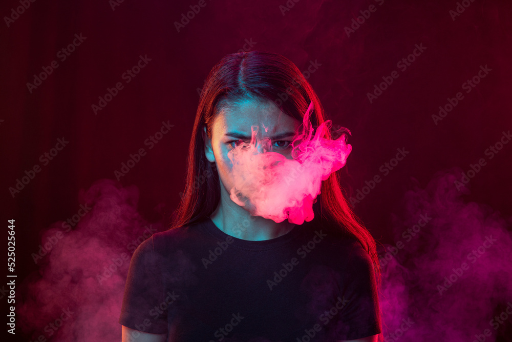 Angry young girl with dark hair releases smoke from the mouth isolated over pink background. Concept of mental health, art, human emotions