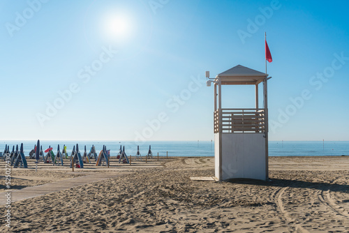 Fototapeta Naklejka Na Ścianę i Meble -  Beach at the Adriatic sea coastline in Italy, Europe during summer. Traditional hut for the beach lifeguard and a red flag.
