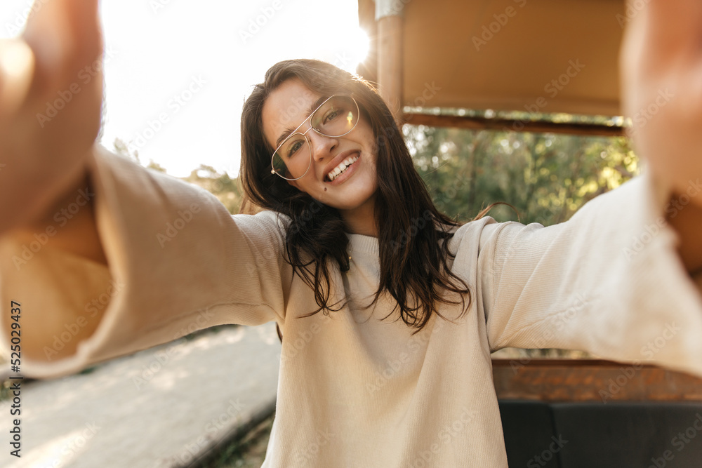 Smiling young caucasian girl takes selfie stretching hands to camera in sunny weather outdoors. Brunette wear glasses, everyday sweatshirt. Emotion concept