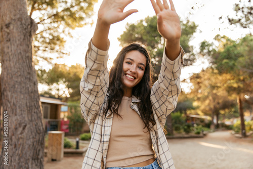 Happy young caucasian woman smiles broadly, stretches out her arms enjoying walk outdoors. Brunette wears t-shirt, shirt and jeans. Weekend concept.