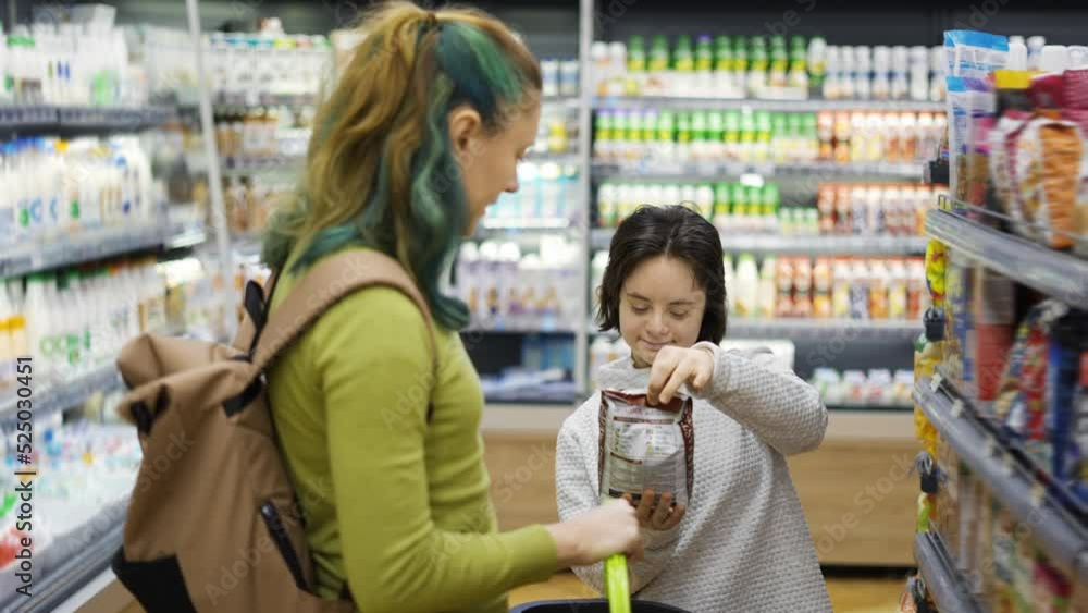 Down syndrome cheerful girl with her mother taking goods from shelf in supermarket, side view Down syndrome cheerful girl with her mother taking goods from shelf in supermarket, side view