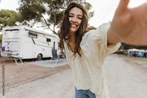 Beautiful young caucasian woman taking selfie holding out her hand to camera outdoors. Brunette wears white blouse and jeans. People emotions, lifestyle and fashion concept.