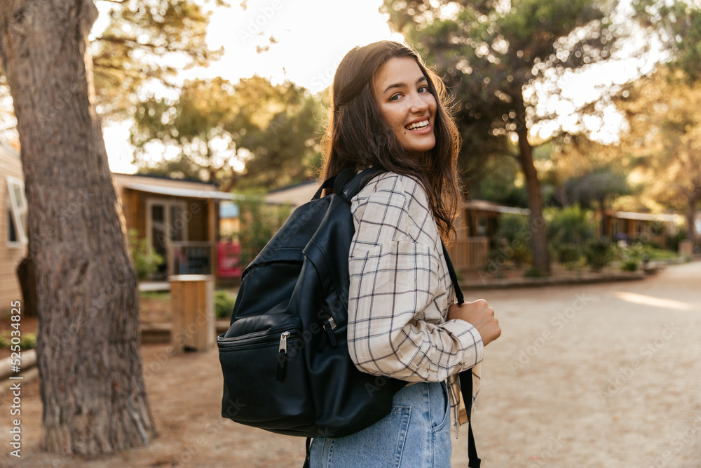Smiling young caucasian girl turns around looking at camera walking on warm spring day on street. Brunette wears shirt, backpack and jeans. Vacation, happiness and pleasure concept