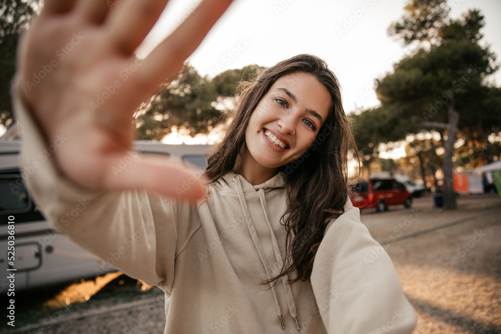 Fun young caucasian girl stretches her palm to camera enjoying spending time outdoors. Brunette smiles broadly, wears beige hoodie. Mood, lifestyle, concept.
