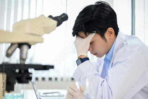 Stressed Asian male scientist or medical technician in white gown at his office desk