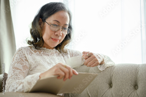 Peaceful Asian middle-aged woman in glasses sits on a sofa reading a book in her living room.