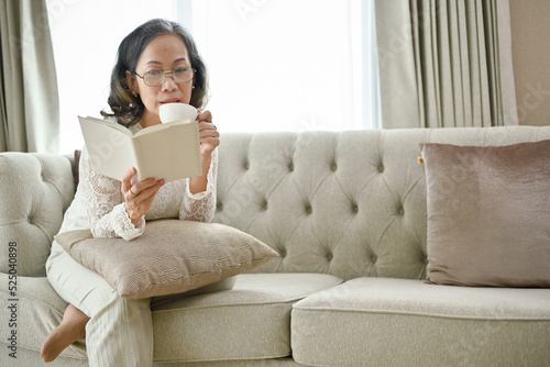 Beautiful Asian-aged woman sipping morning coffee while reading a book in her living room.
